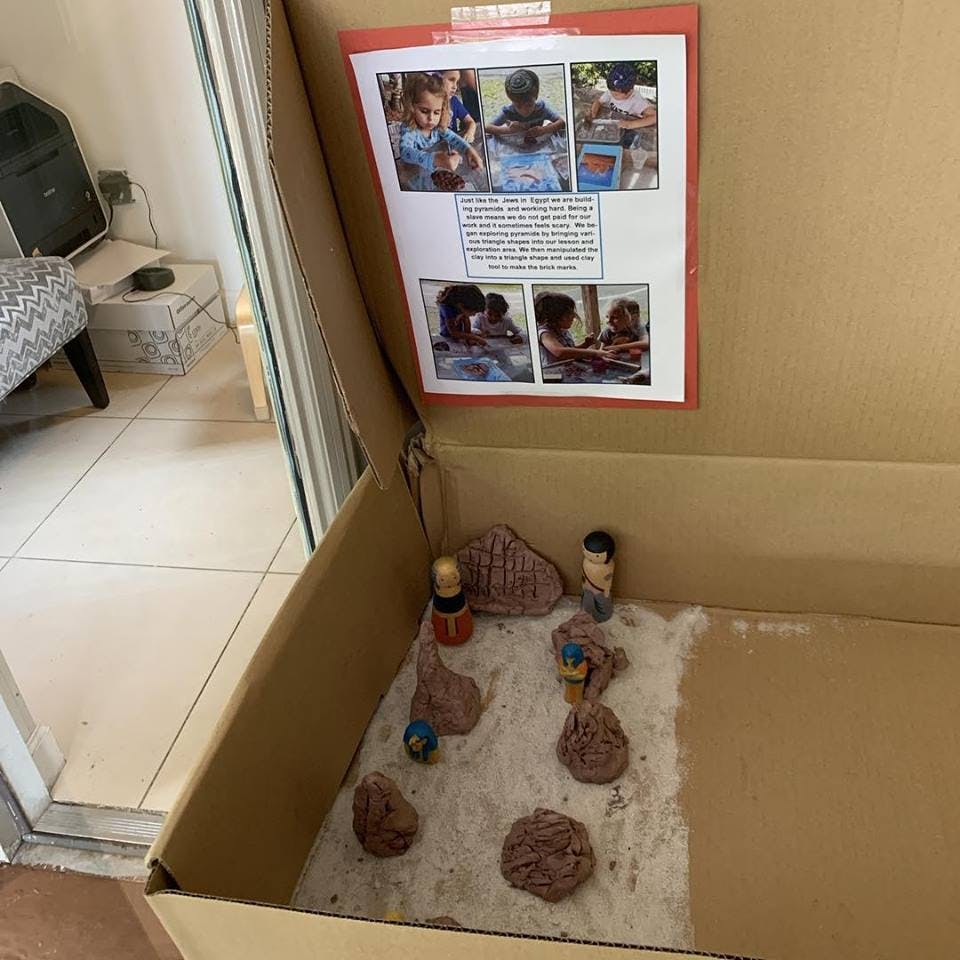 Cardboard box with sand, rocks, and small figures, with a paper showing photos of children