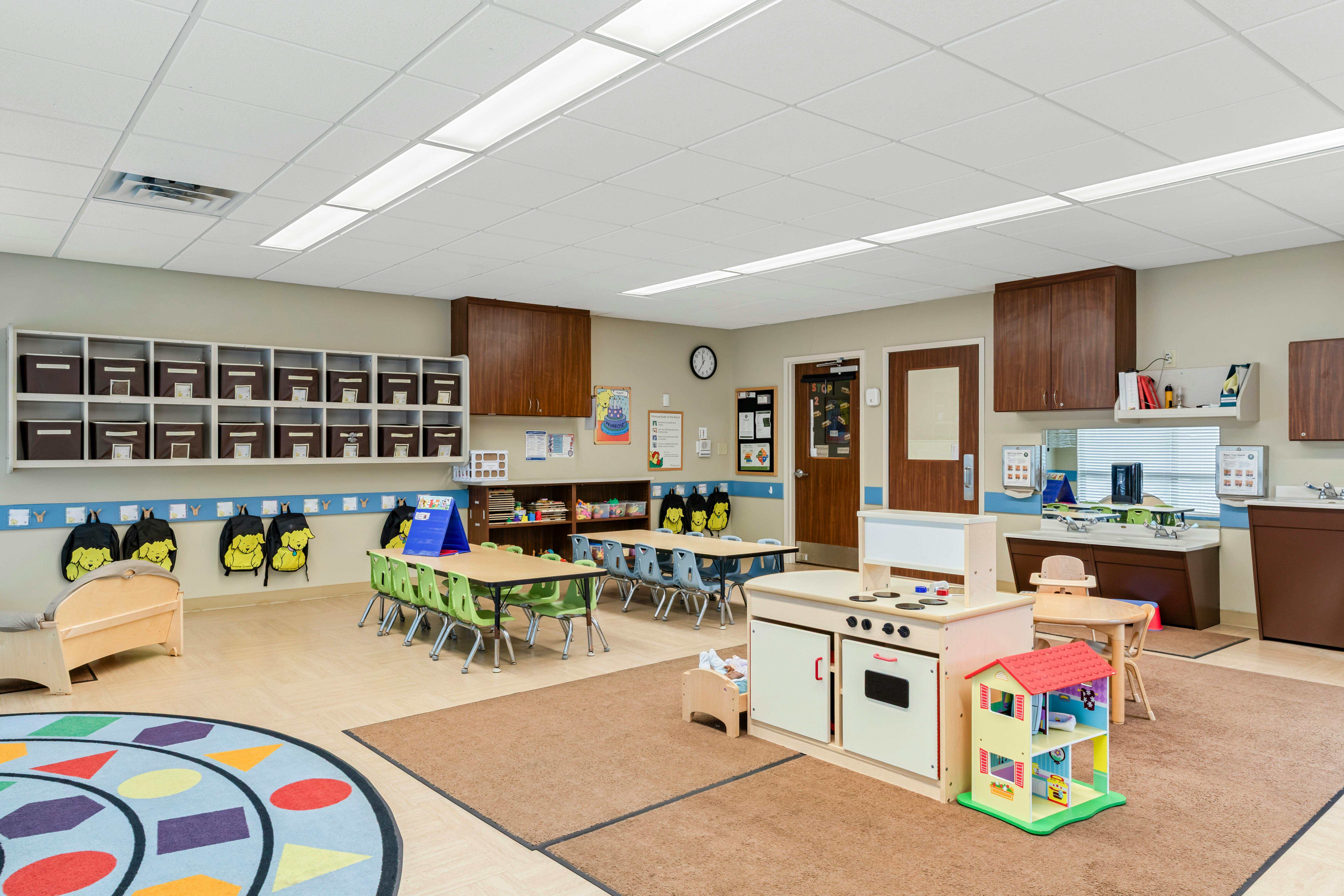 Empty classroom with tables, chairs, cubbies, play kitchen, and a circular rug
