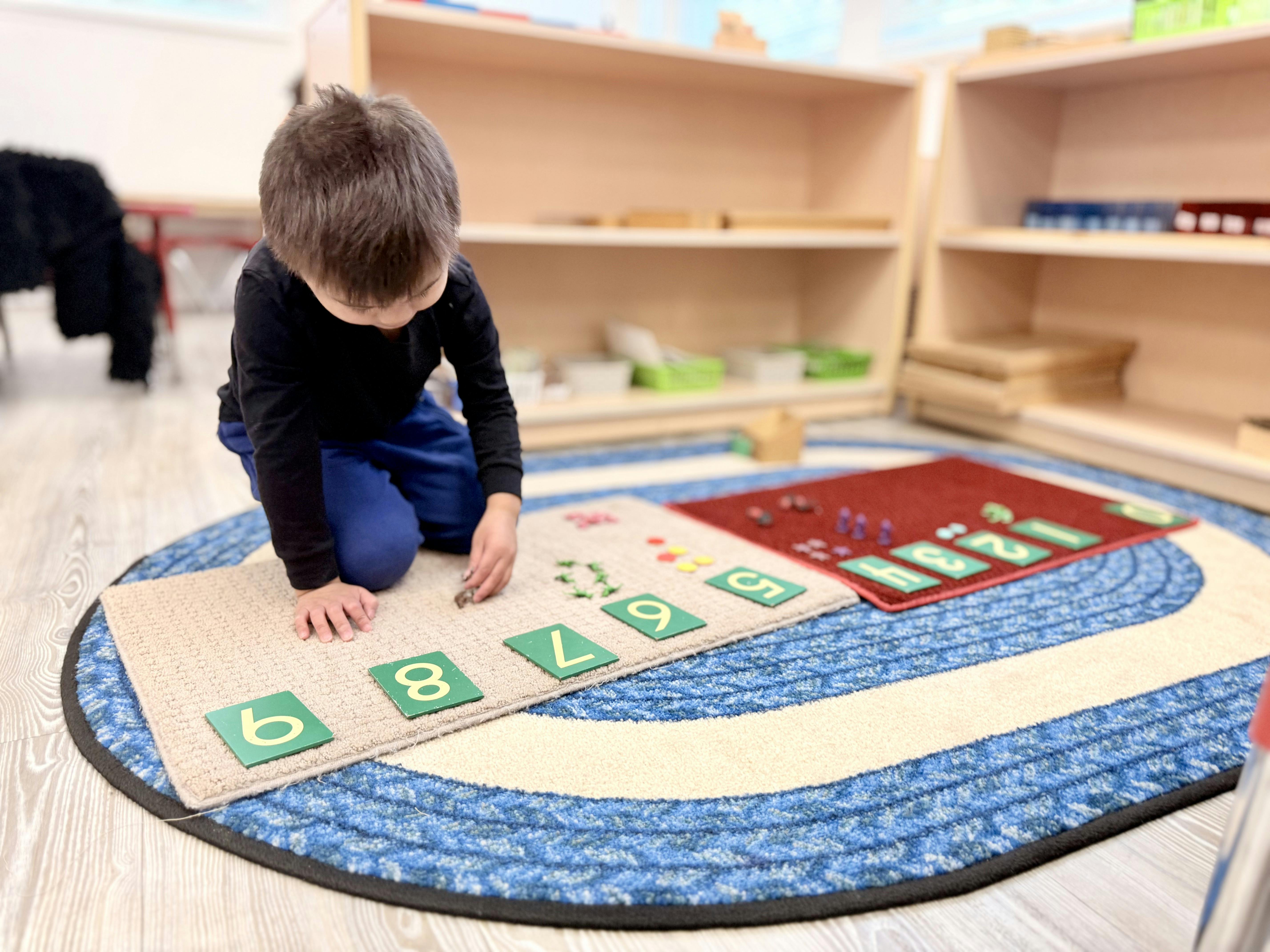 Child kneeling on a rug, placing number tiles on a mat in a classroom