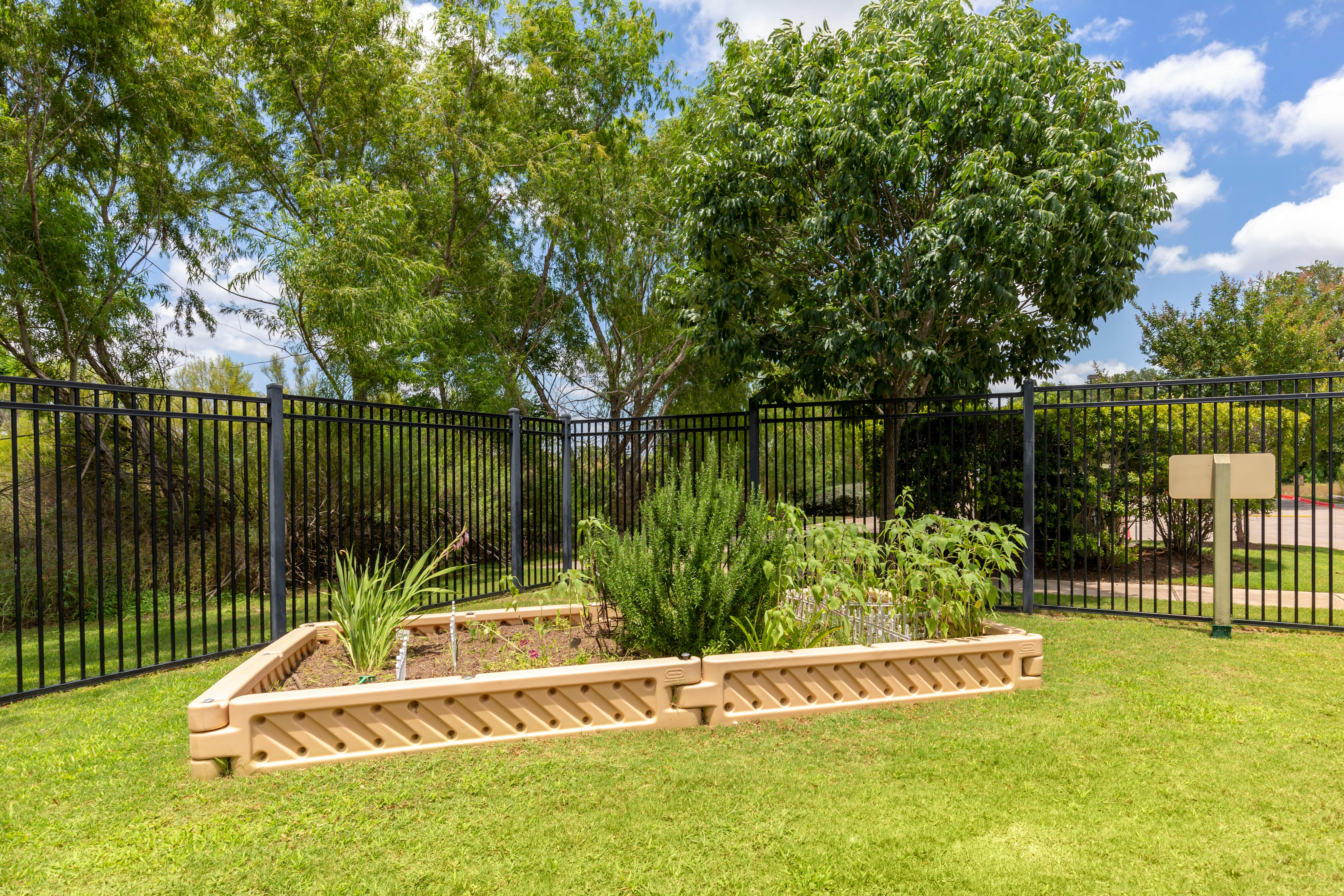 A beige raised garden bed with various plants on a grassy area, next to a black fence
