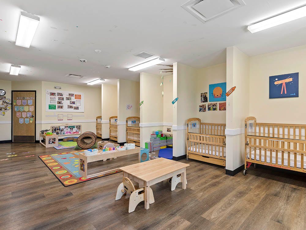 A daycare room with several wooden cribs, a play rug, and small tables with chairs