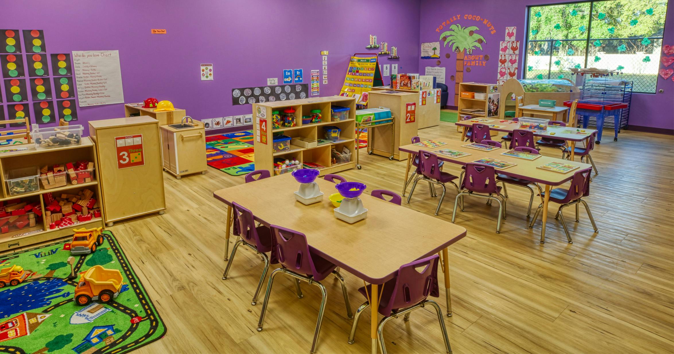 A colorful classroom with small tables and chairs, shelves with toys, and activity charts on the walls