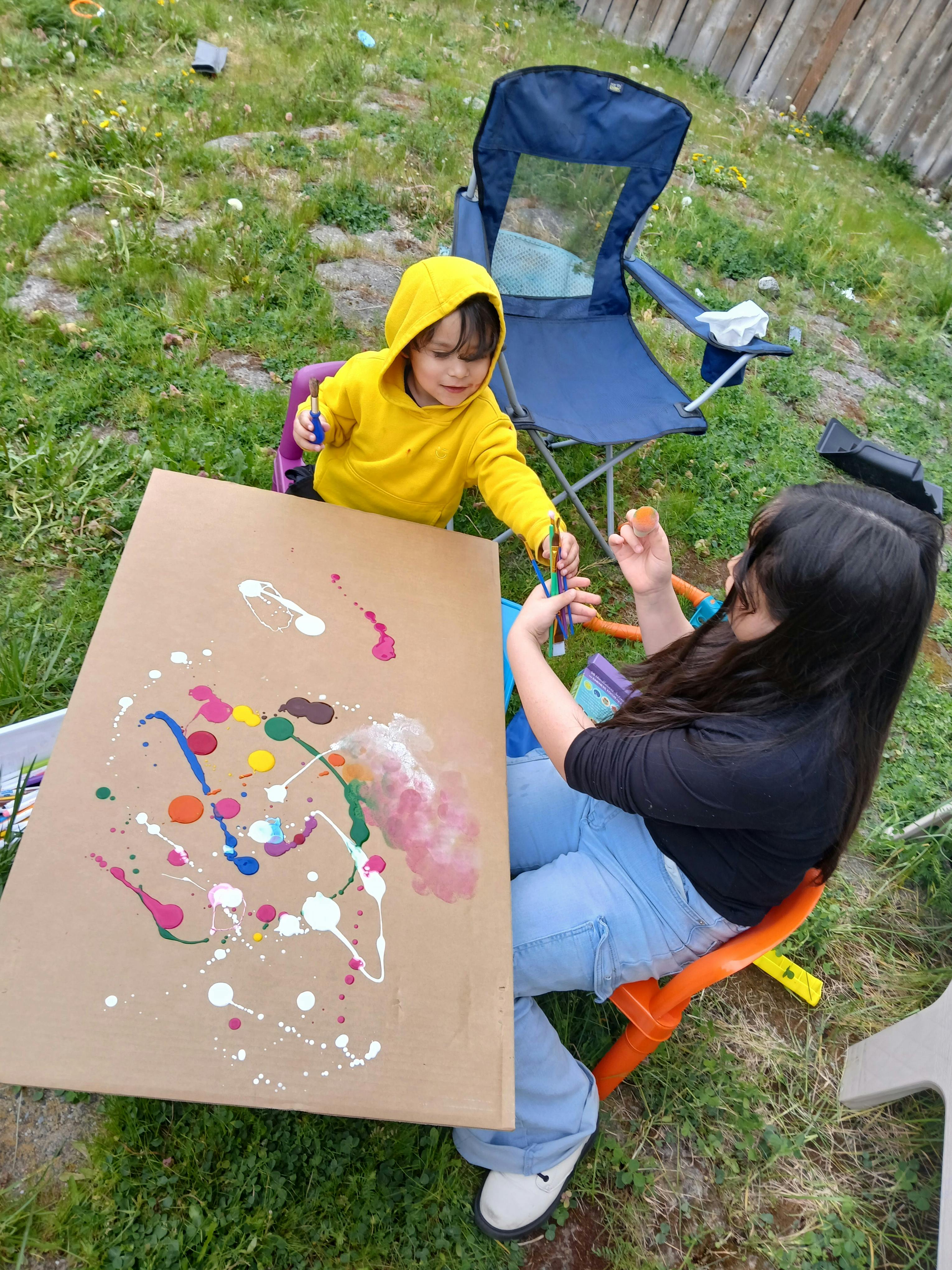 A person helping a child paint on a large piece of cardboard outdoors