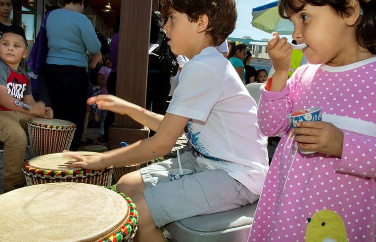 Child playing a drum next to another child eating from a cup