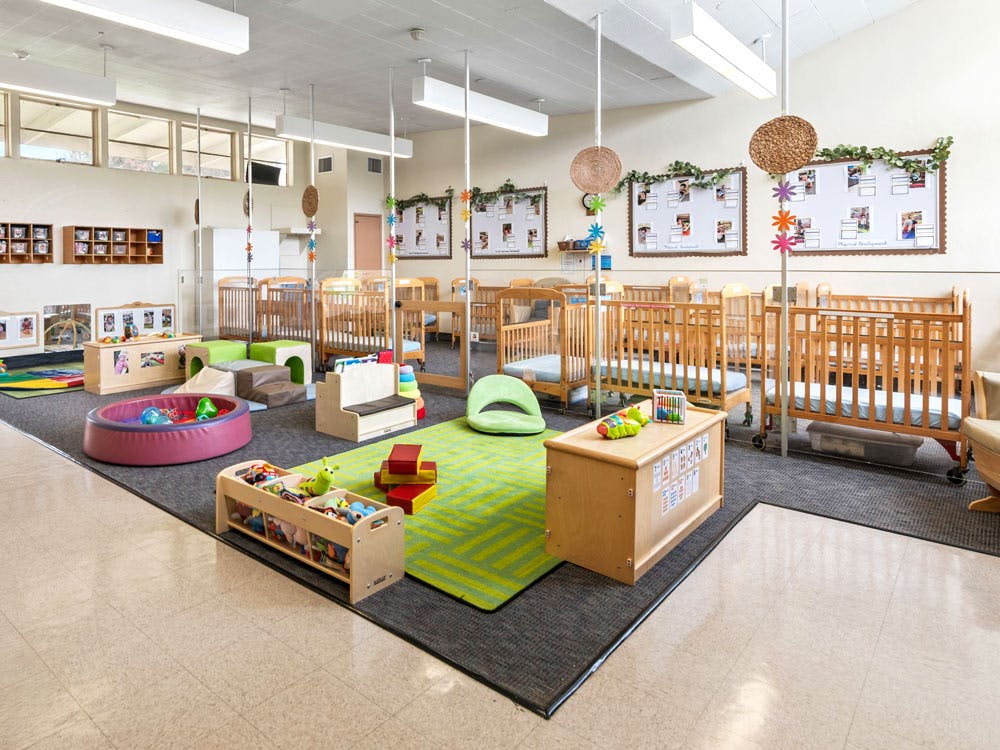 A brightly lit classroom with multiple cribs, soft play equipment, and toys