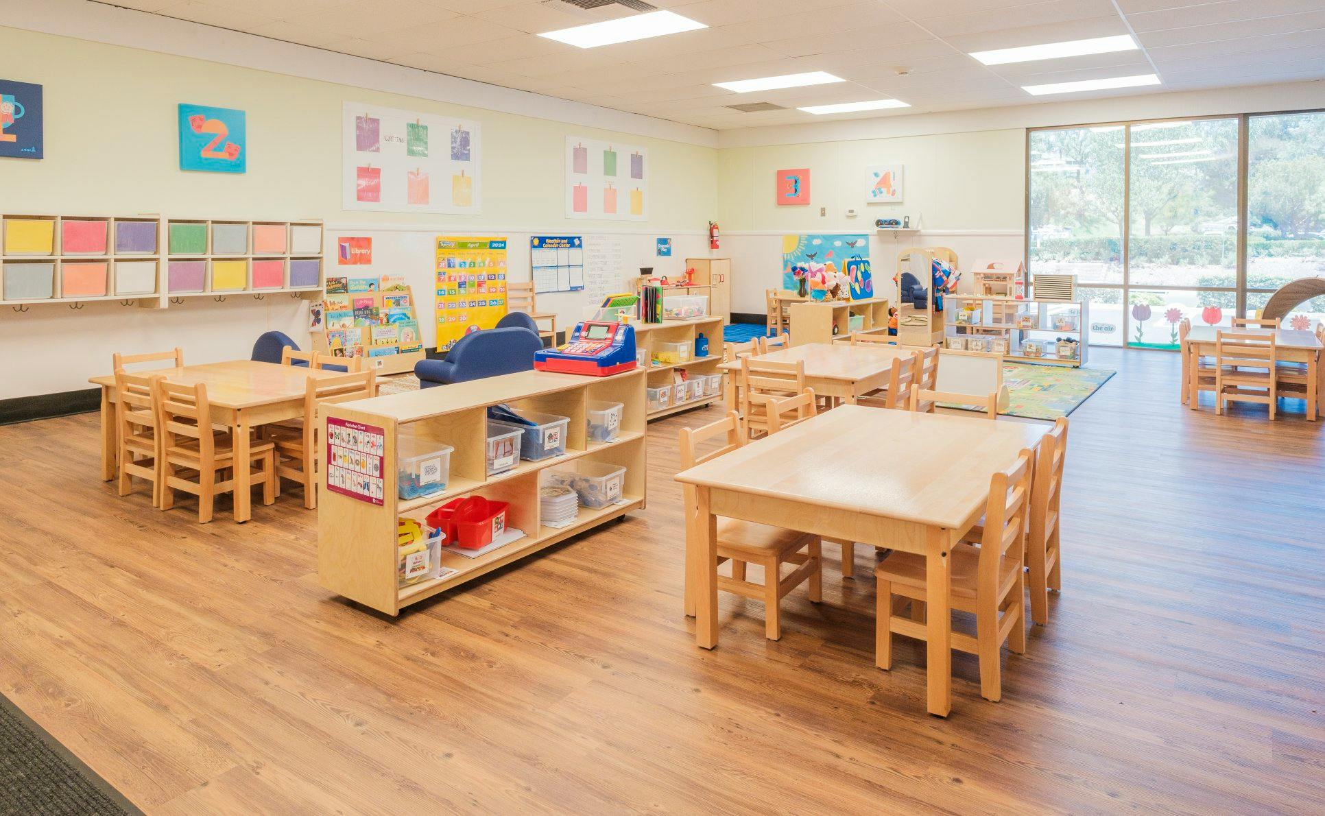 An empty classroom with small wooden tables and chairs, and shelving units