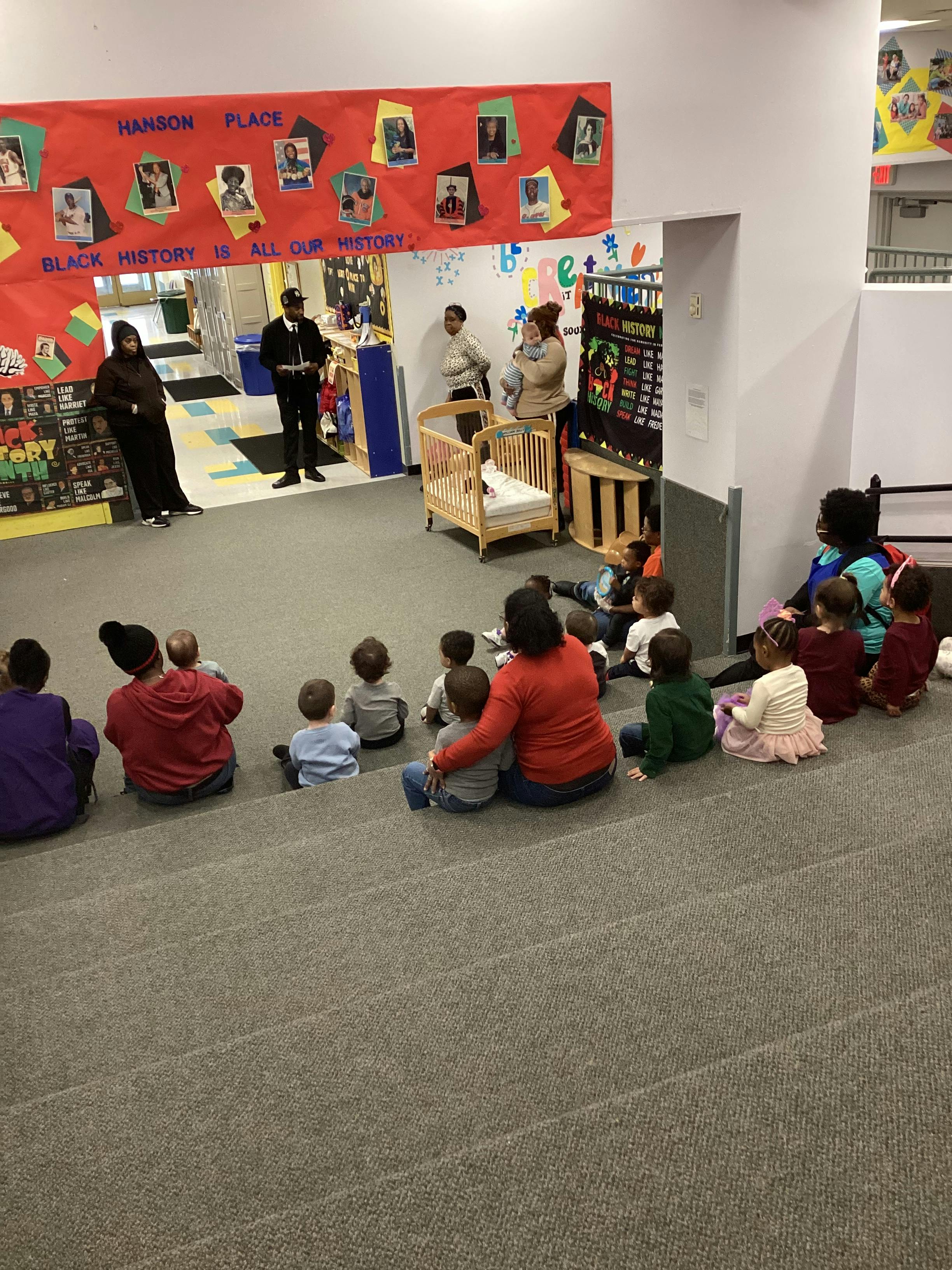 Adults and children gathered on the floor around two standing adults in a classroom