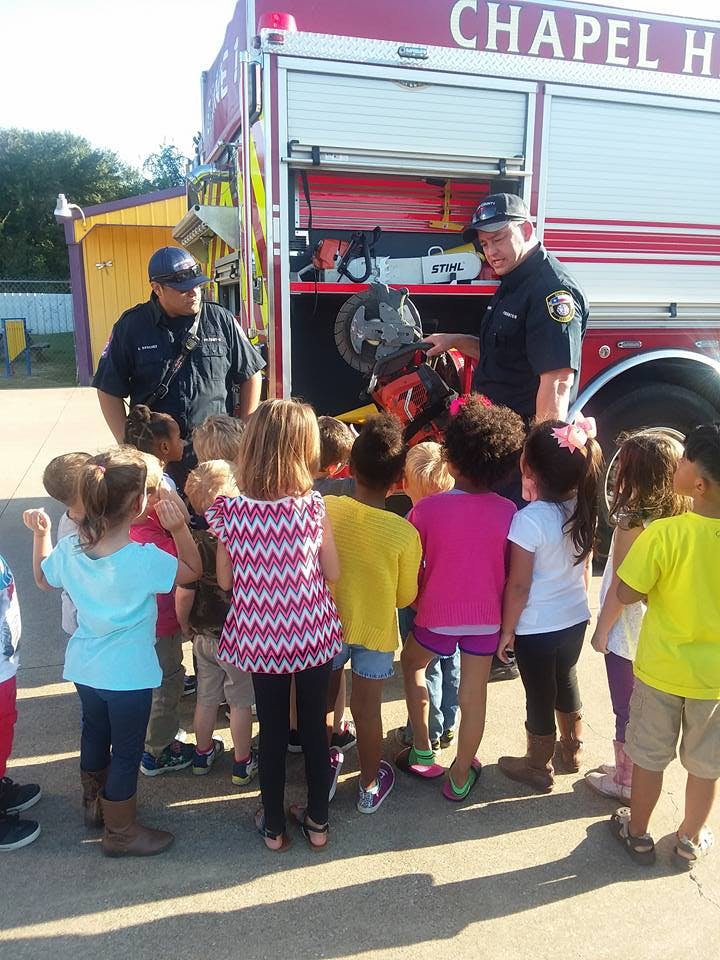Two people in uniform showing equipment to a group of children near a fire truck
