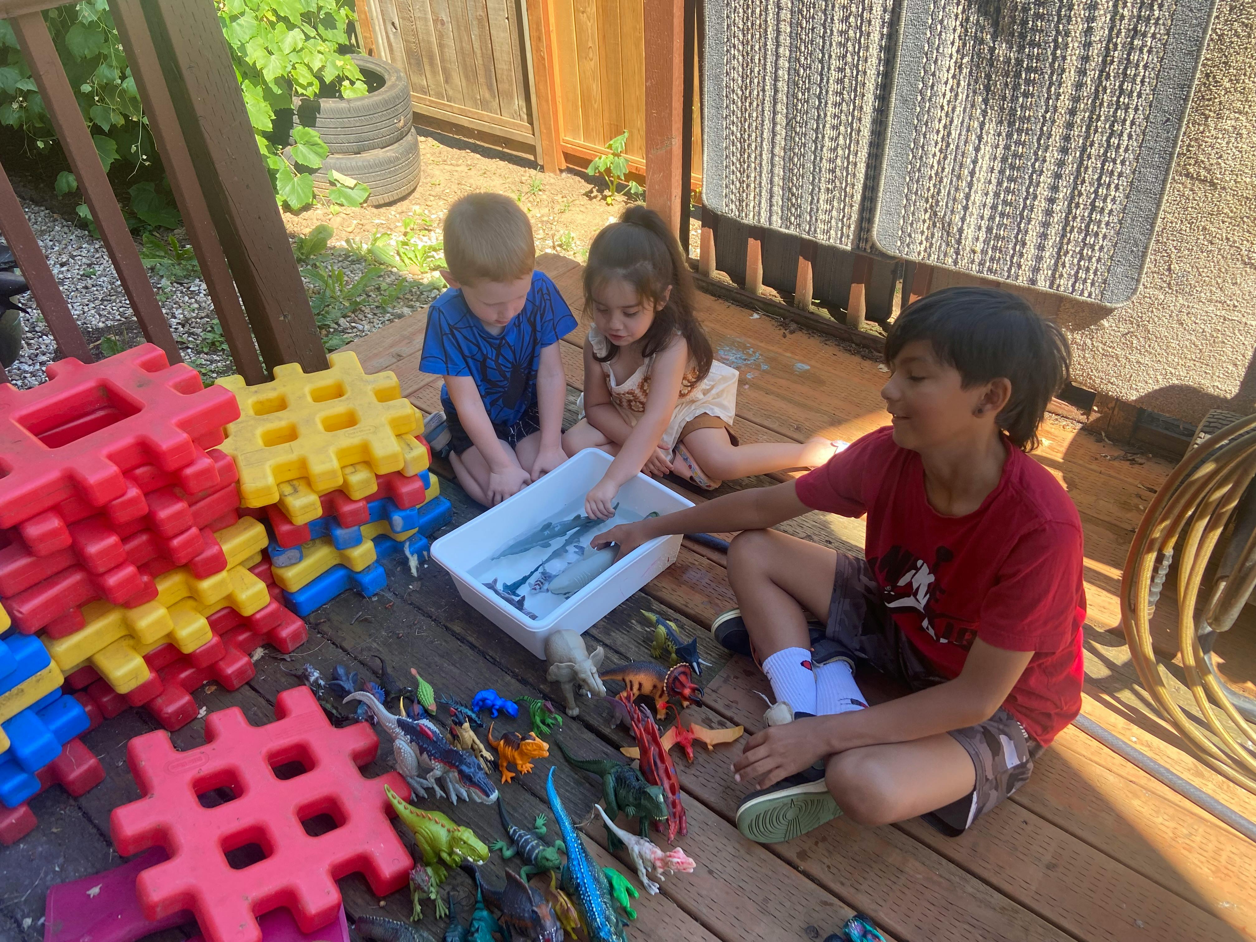 Three children playing with toys and a tub of water on a wooden deck
