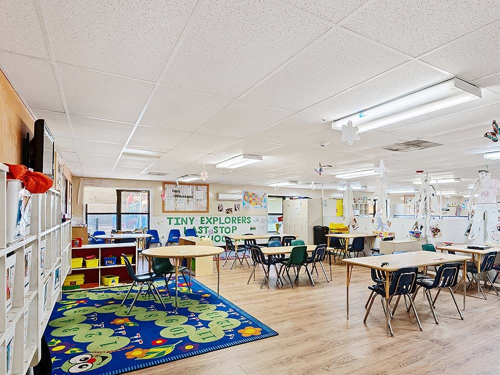 Empty classroom with small tables and chairs, a play rug, and shelves