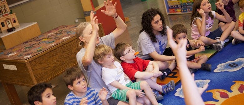 Adults and children sitting on a rug, some with hands raised