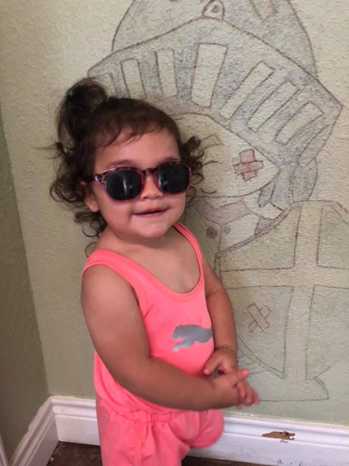 Child wearing sunglasses and a pink outfit, standing next to a wall drawing