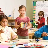 Child holding a box of crayons in a classroom with other children