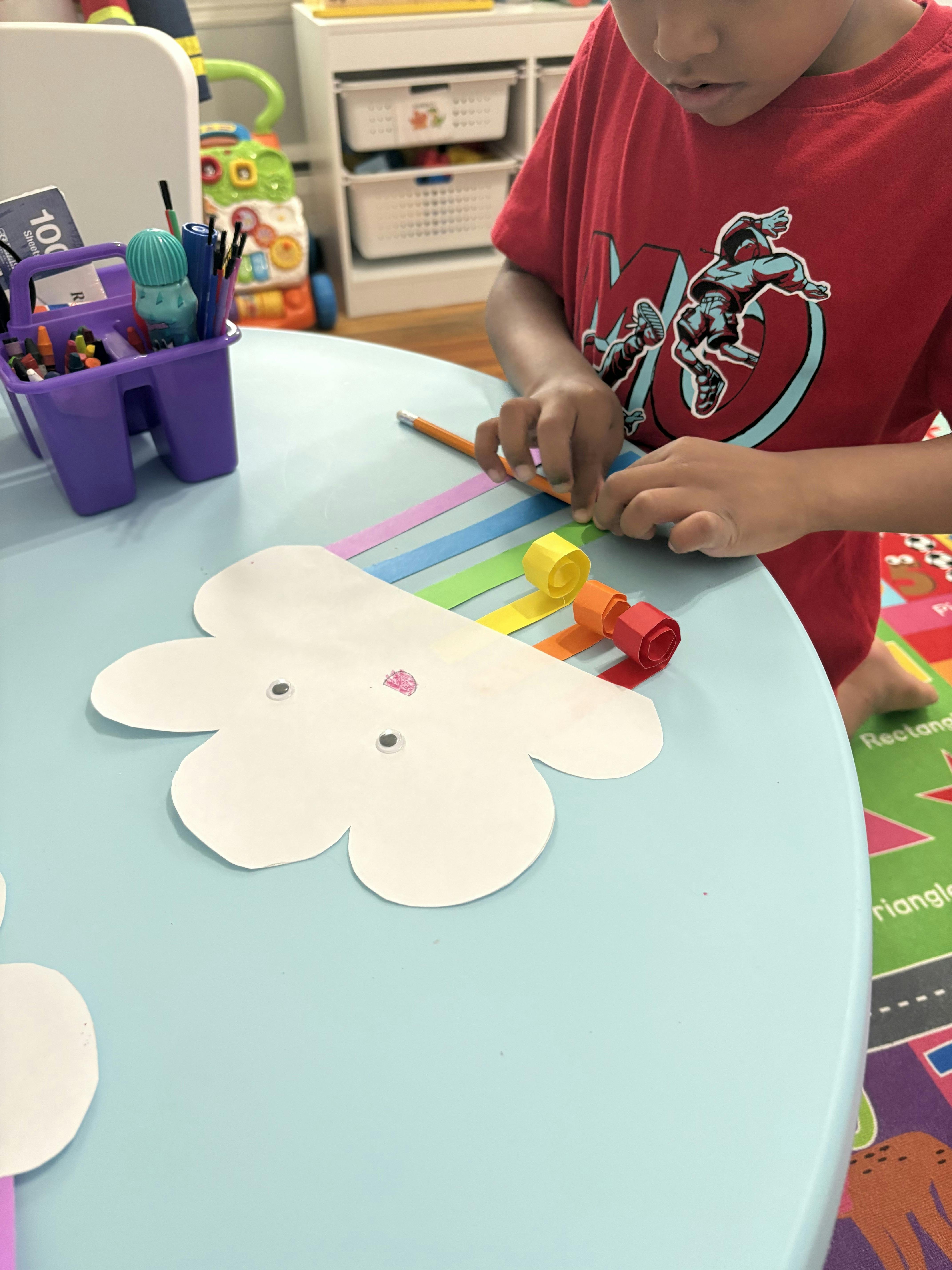 Child crafting a paper cloud with curled rainbow strips at a table