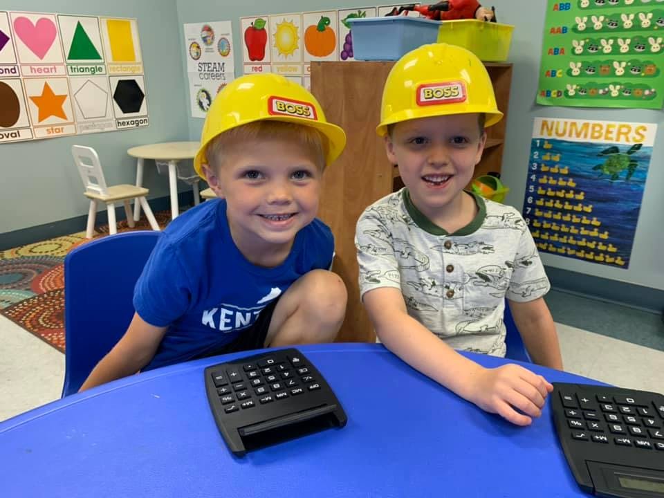 Two children wearing yellow hard hats sitting at a table with calculators
