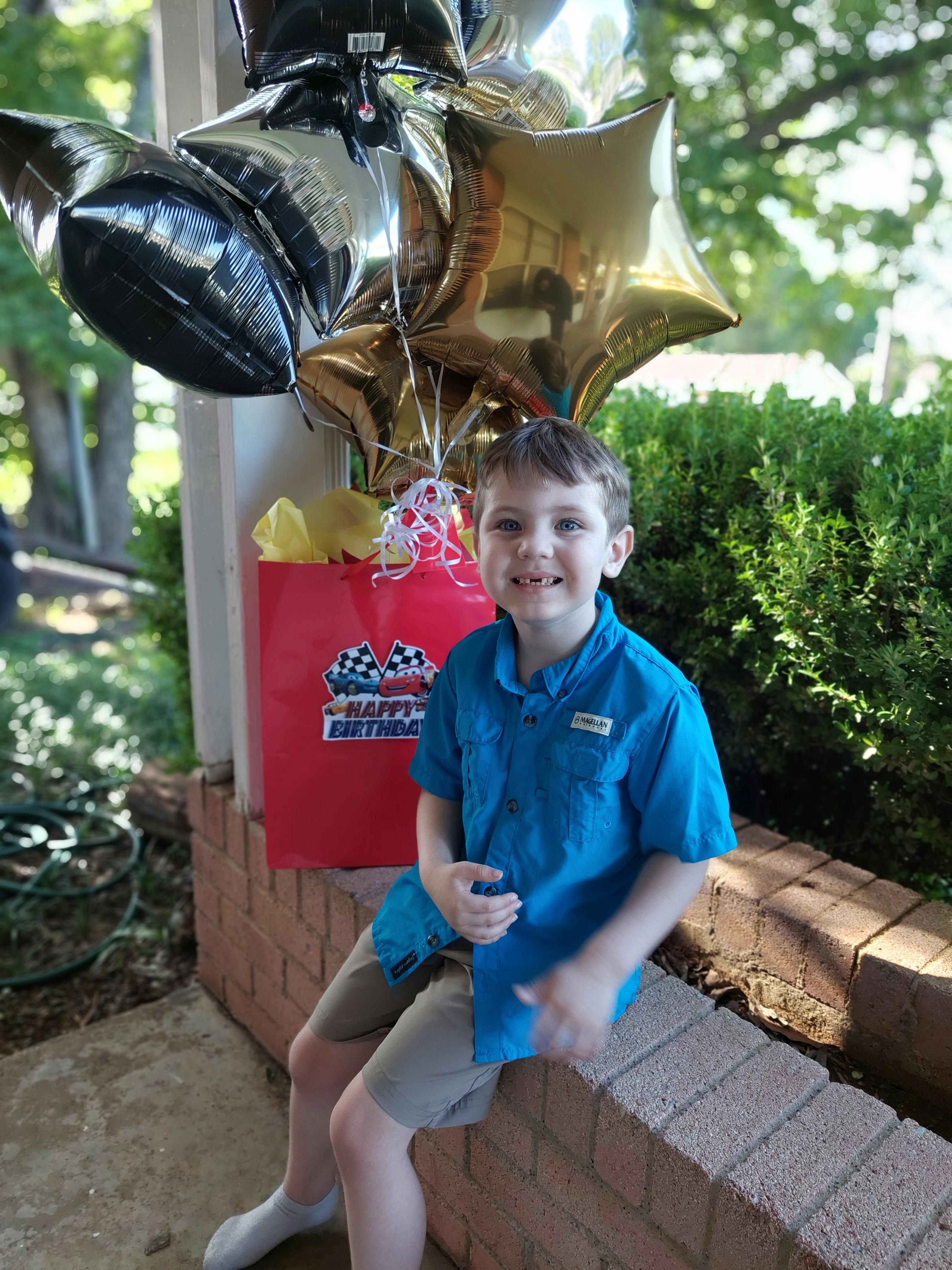 Child sitting on a brick ledge with birthday balloons and a gift bag