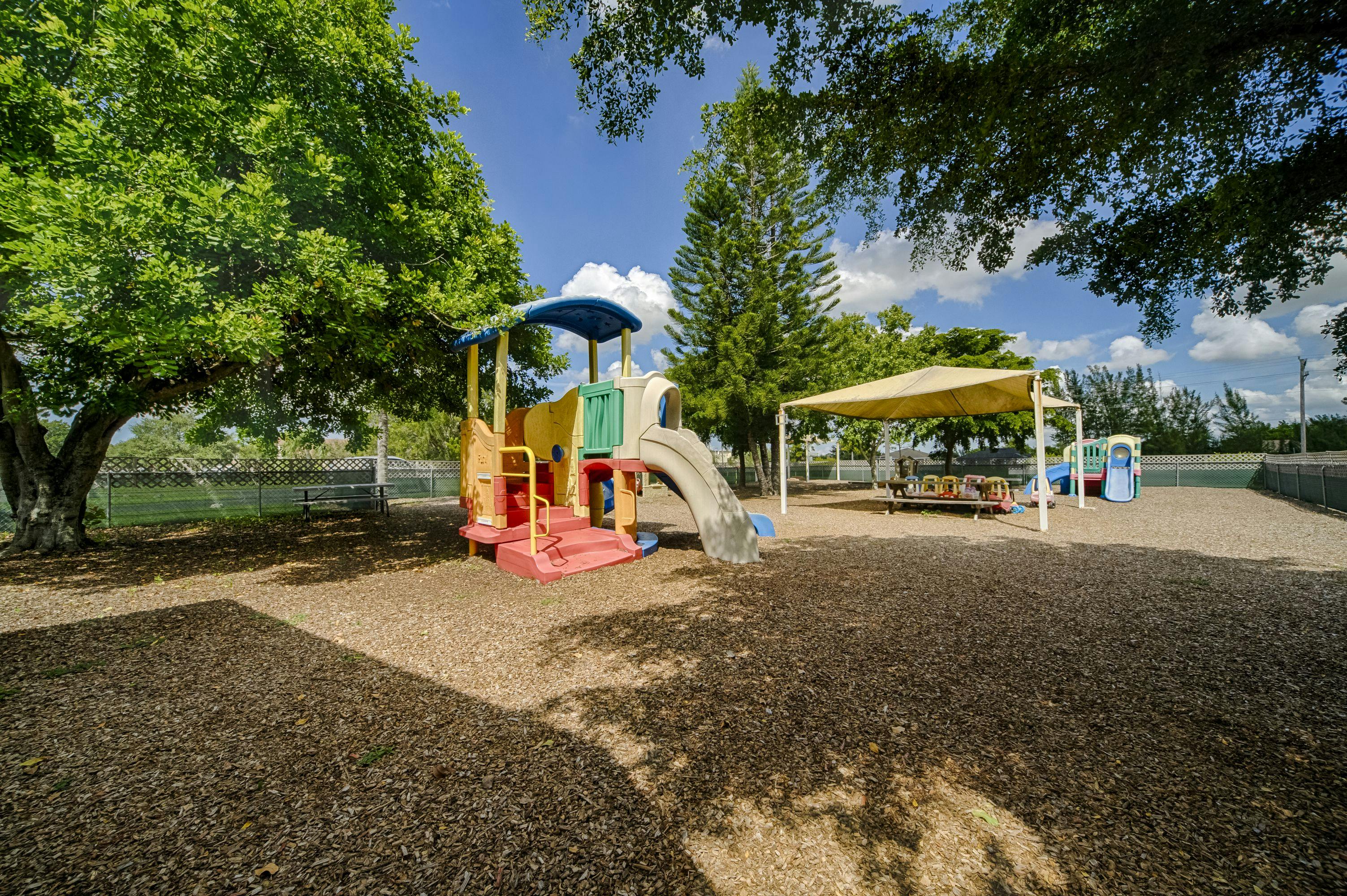 Playground with a slide and climbing structure, shaded by trees and a canopy