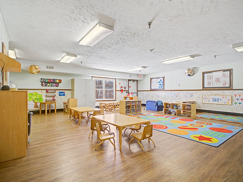 Empty classroom with small tables and chairs, colorful rugs, and play areas
