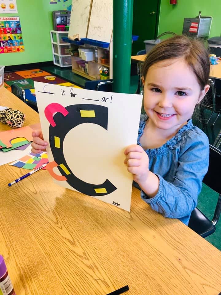 Child holding up a paper with a large letter C and text "C is for __ar!"