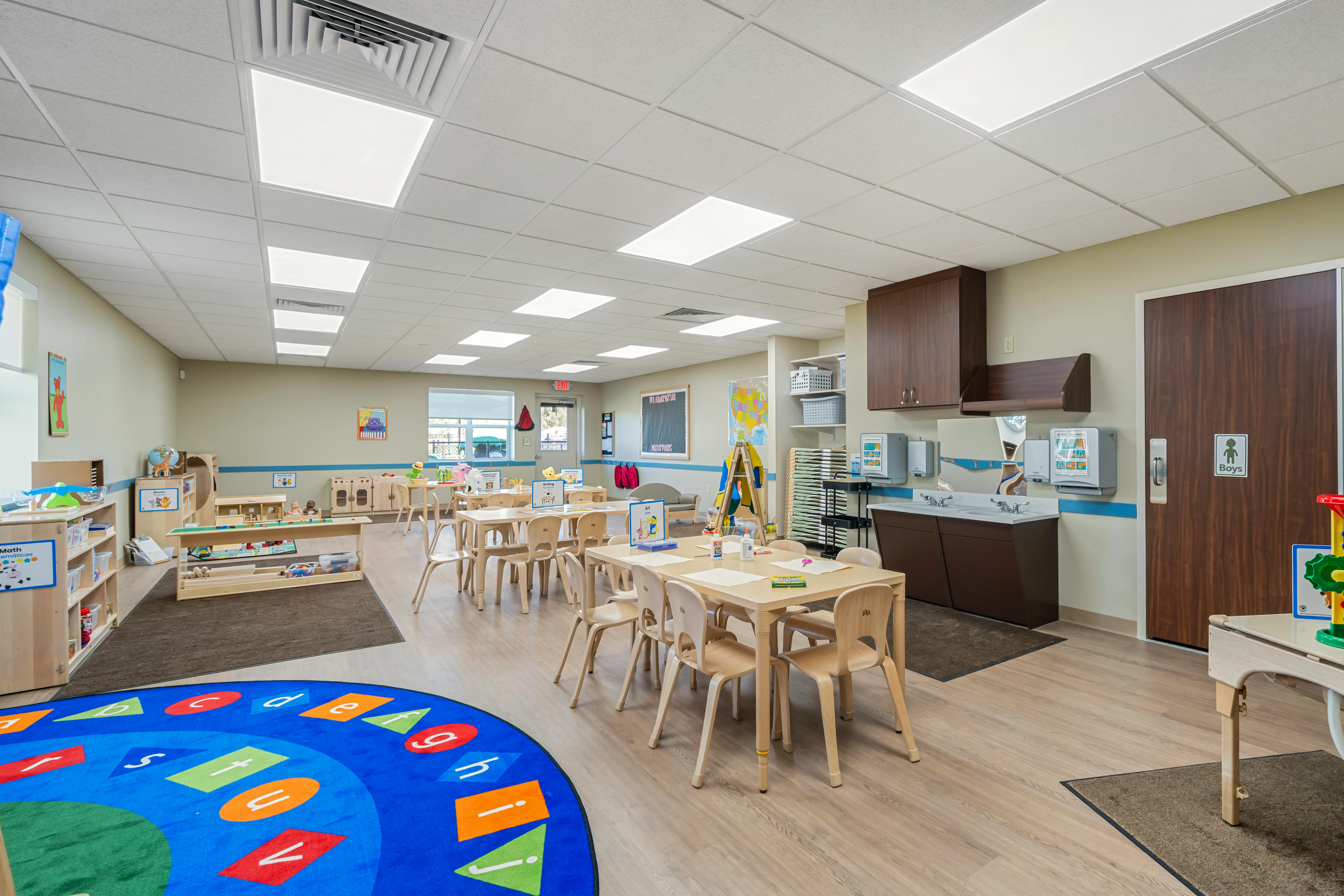 An empty early childhood classroom with small tables and chairs, a sink, and a colorful rug