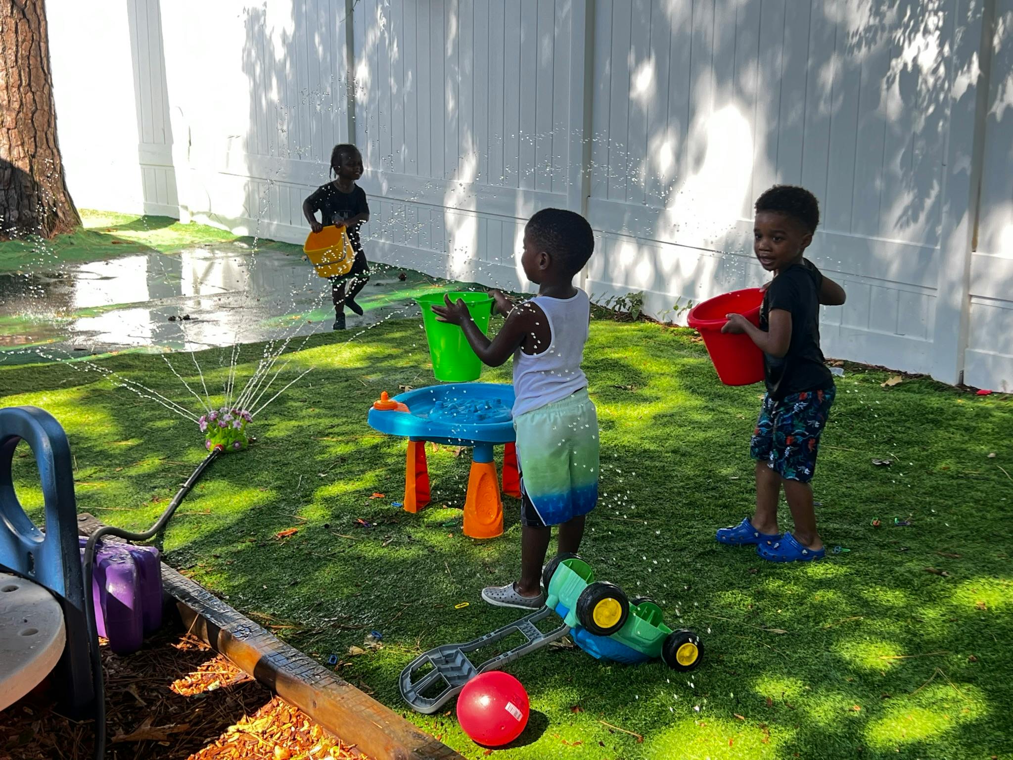 Three children playing with buckets and a sprinkler in a grassy area