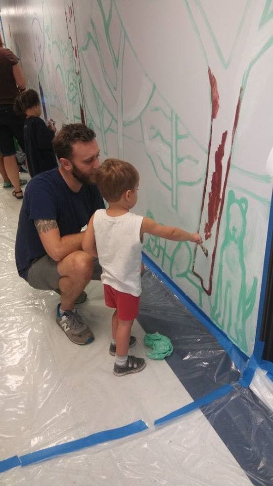 Person kneeling next to a child painting on a wall