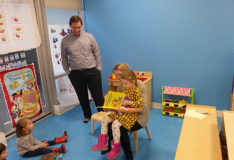 Adult reading a book to a child in a classroom, another child on the floor