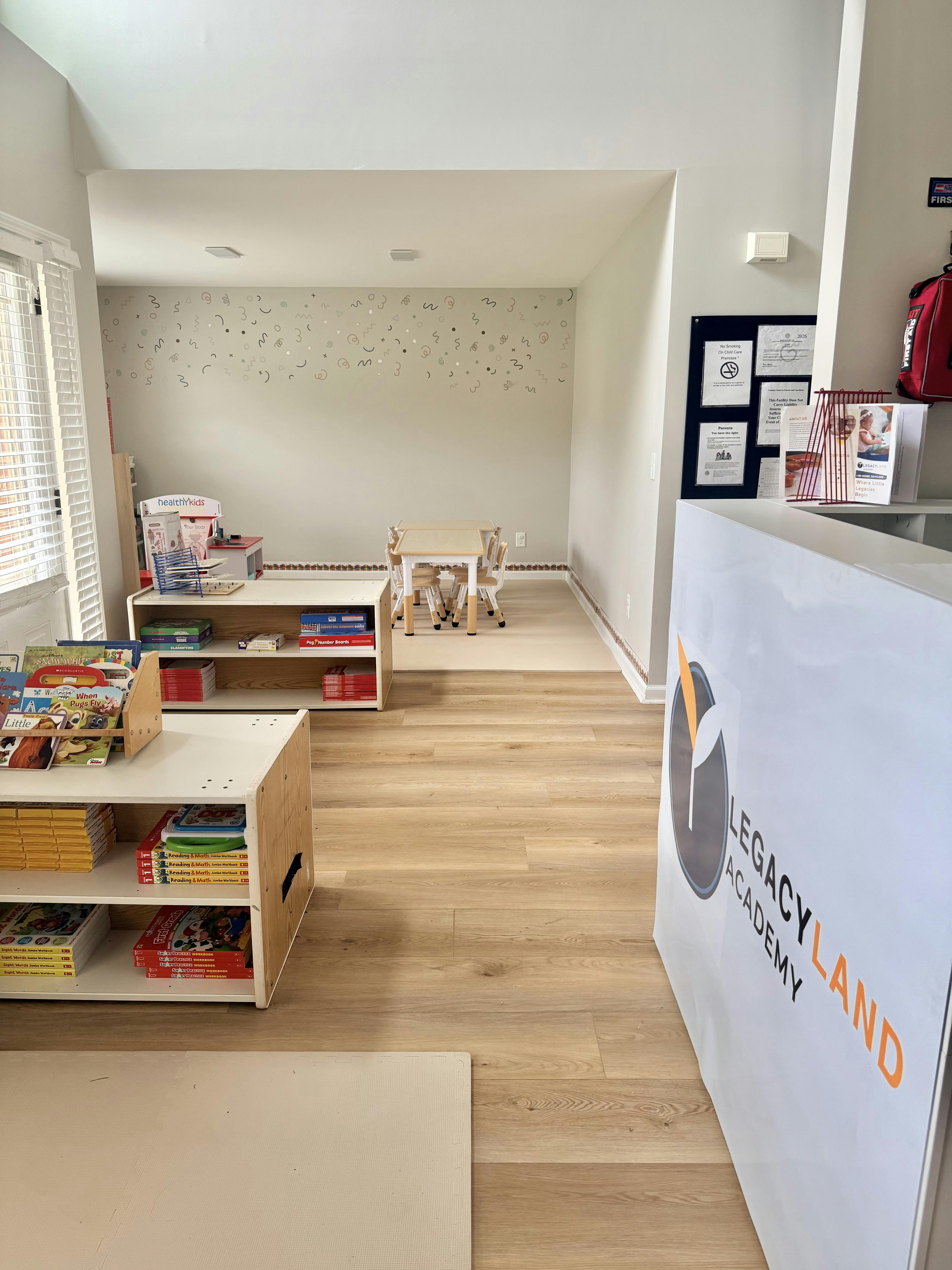 Classroom with child-sized tables and chairs, shelves of books and toys, and a "LEGACY LAND ACADEMY" sign
