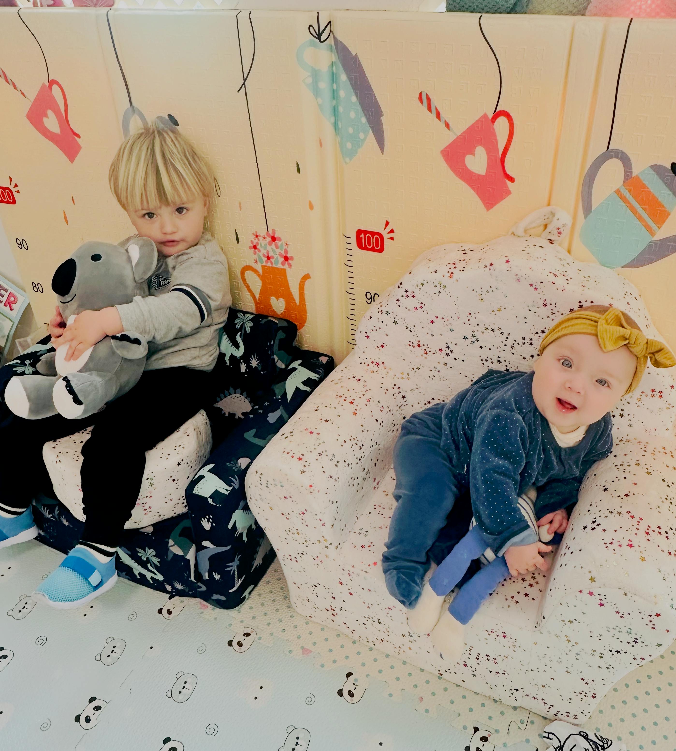 Child holding a koala plush toy and an infant sitting in small chairs