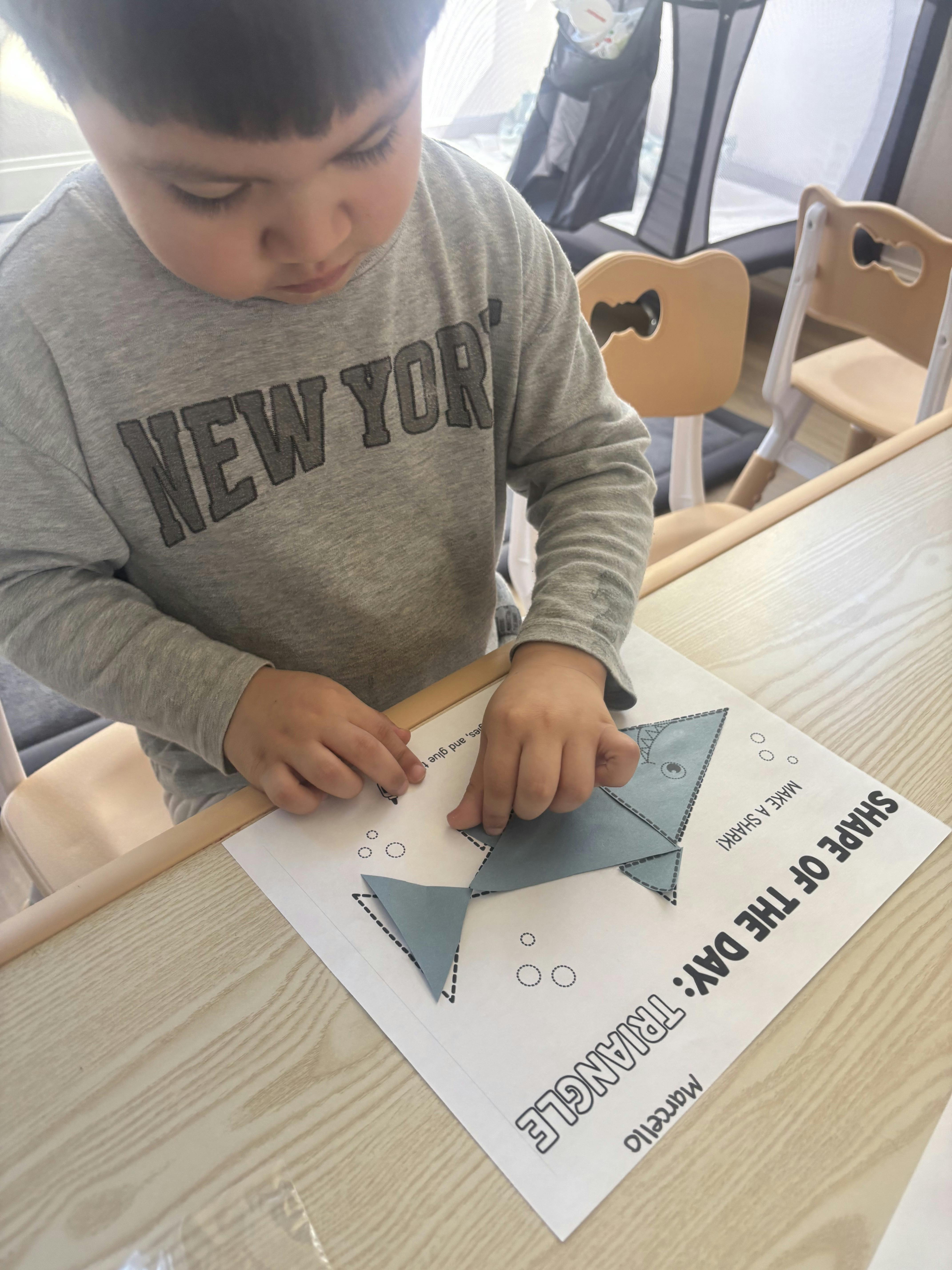 Child assembling a paper shark craft labeled "Shape of the Day: Triangle"