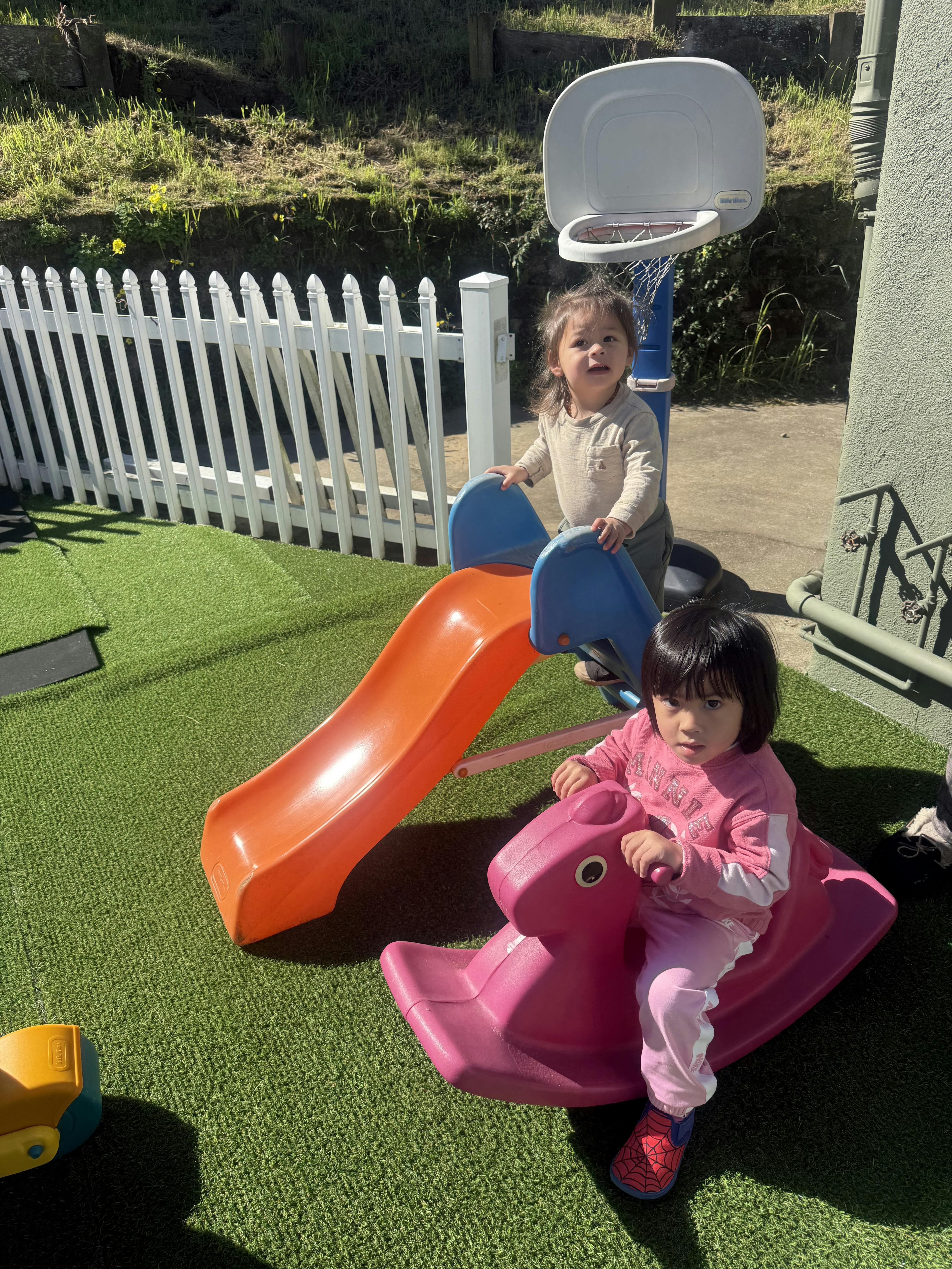 Two children playing on an orange slide and pink rocking horse on artificial grass