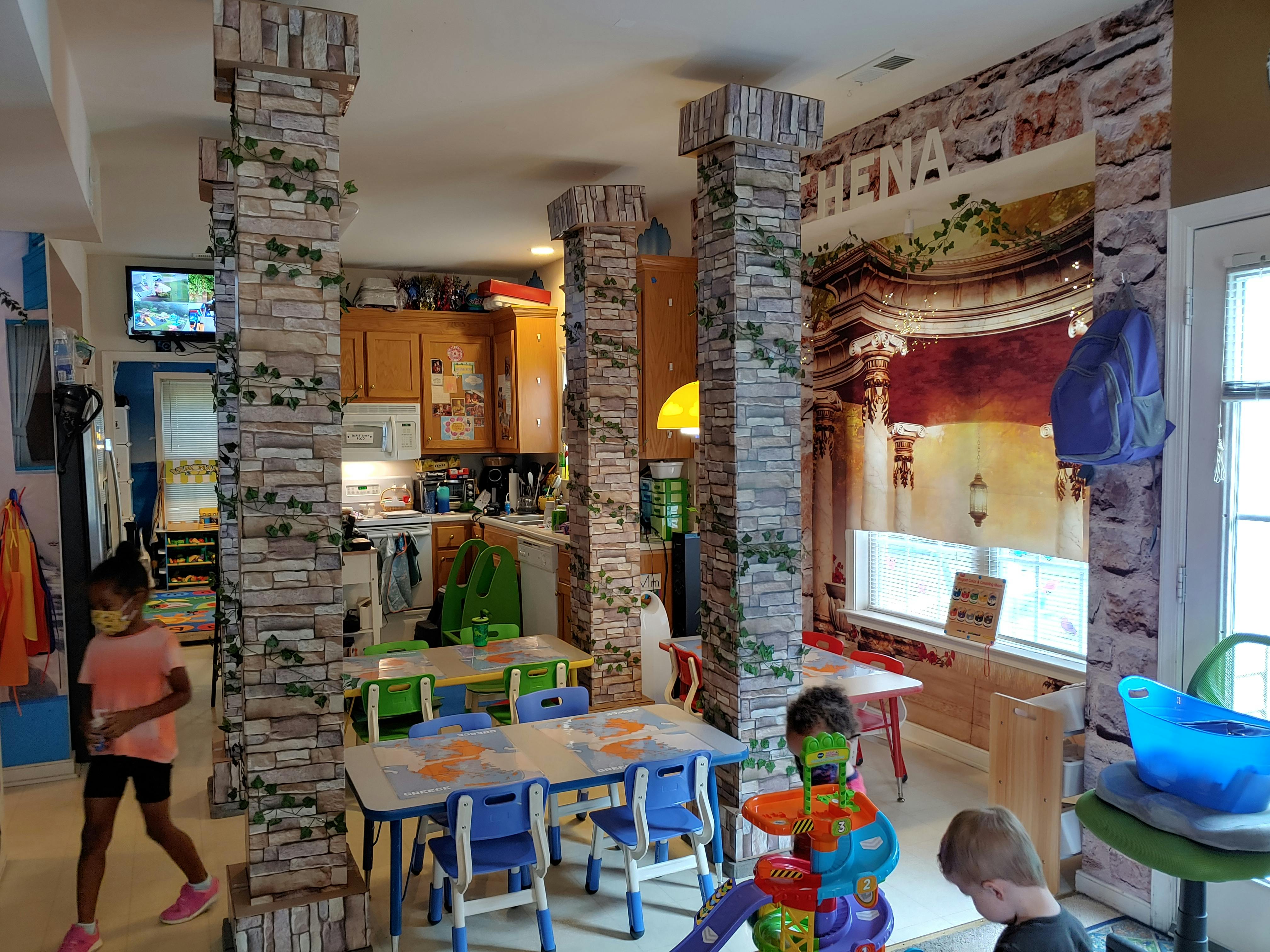 Children in a room with tables and chairs, and a kitchen in the background