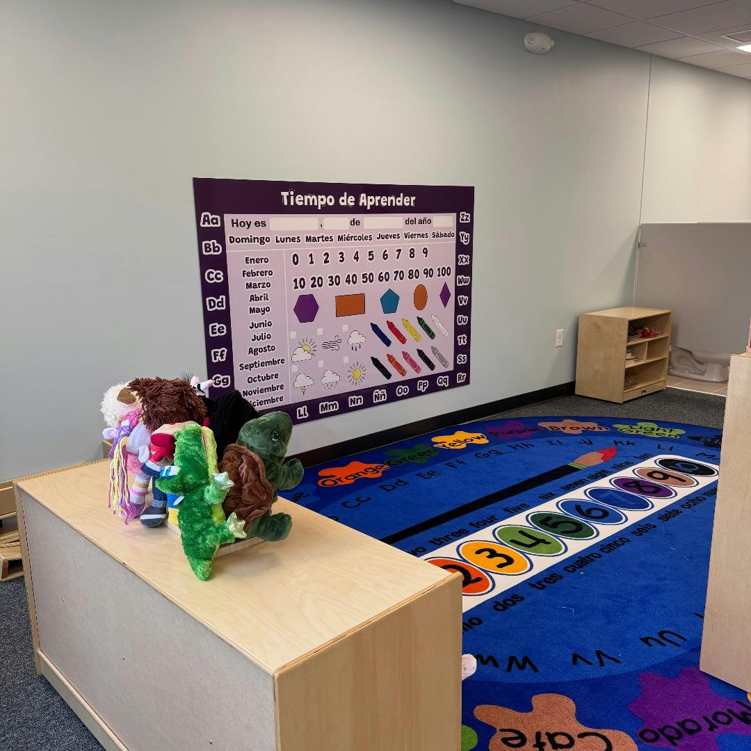 Classroom with a Spanish learning poster, blue rug, and stuffed animals on a wooden shelf
