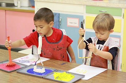 Two children painting with various tools in a classroom