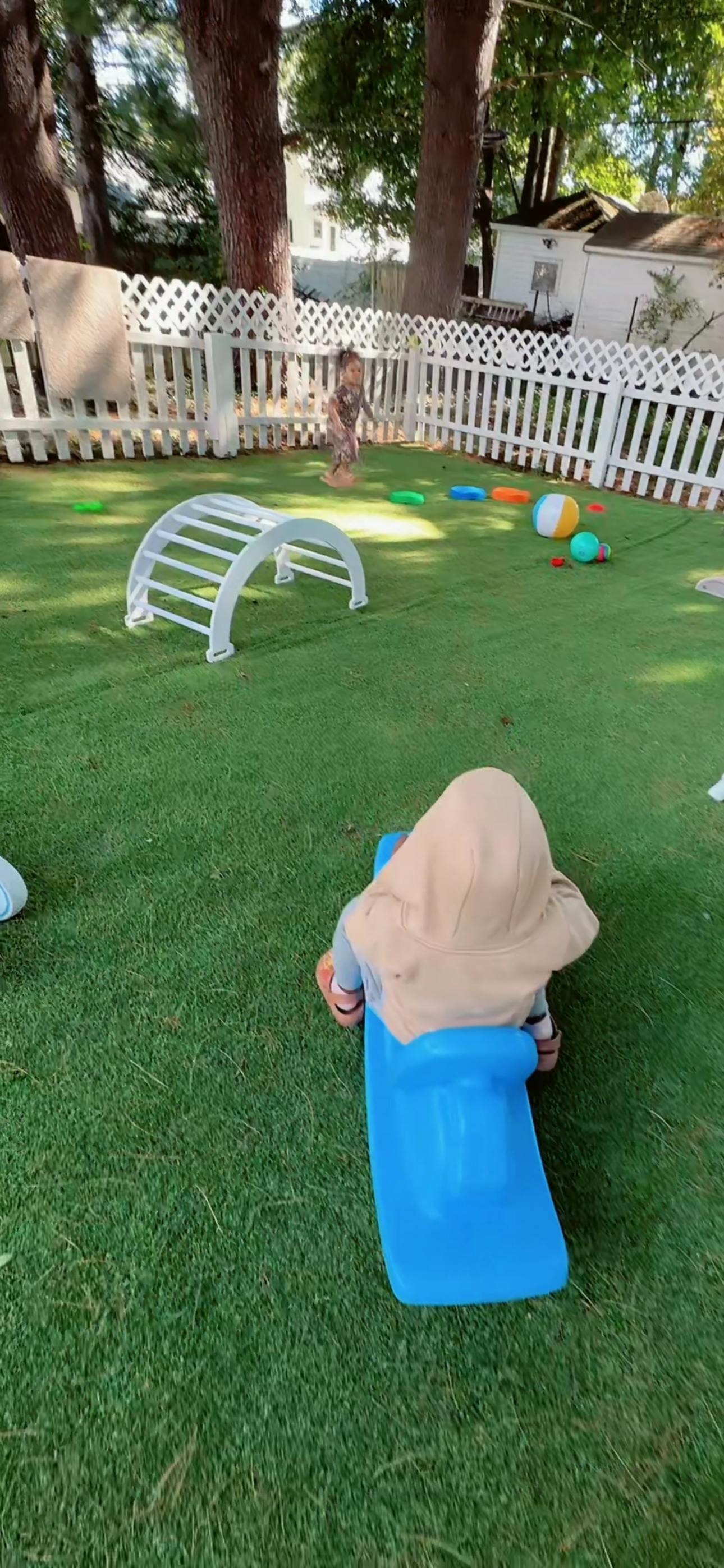 Child on a blue seesaw in a backyard with another child in the background