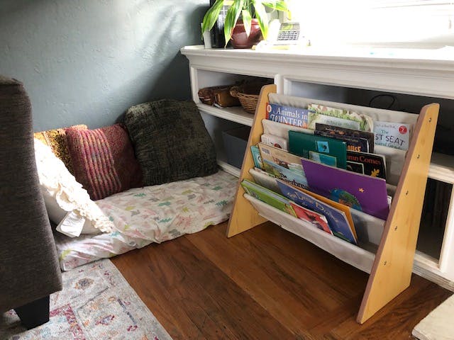 Floor cushion with pillows next to a small wooden bookshelf filled with books