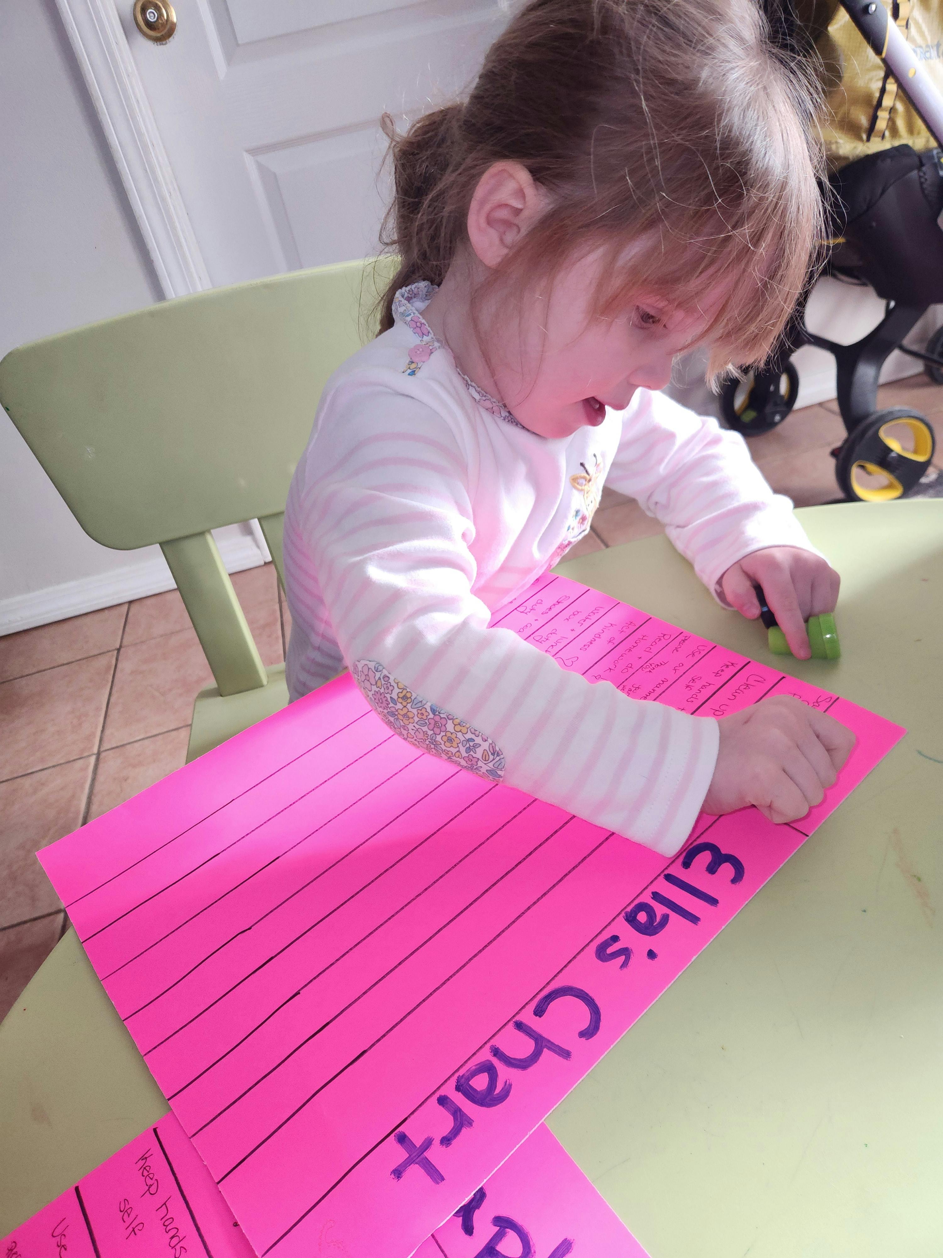 Child writing on a pink paper with "Ella's Chart" written on it