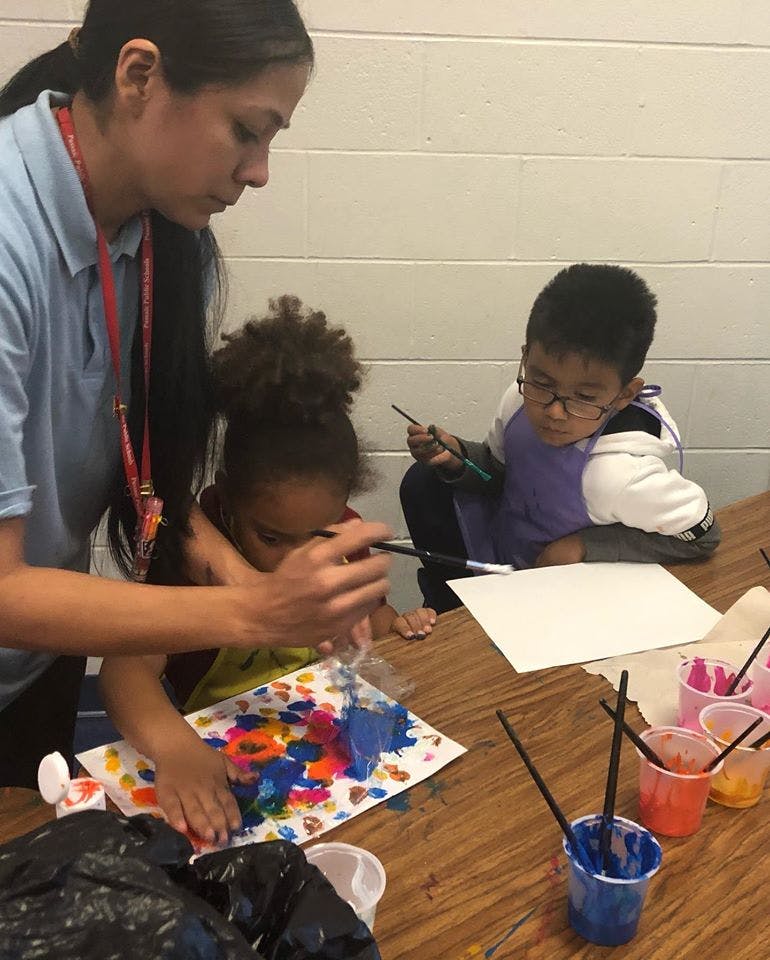 Person helping a child paint, another child painting nearby