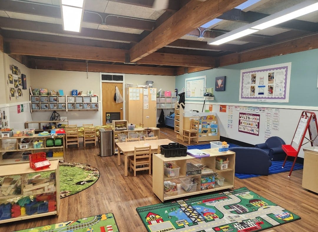 Classroom with wooden tables, chairs, shelving units, and activity rugs