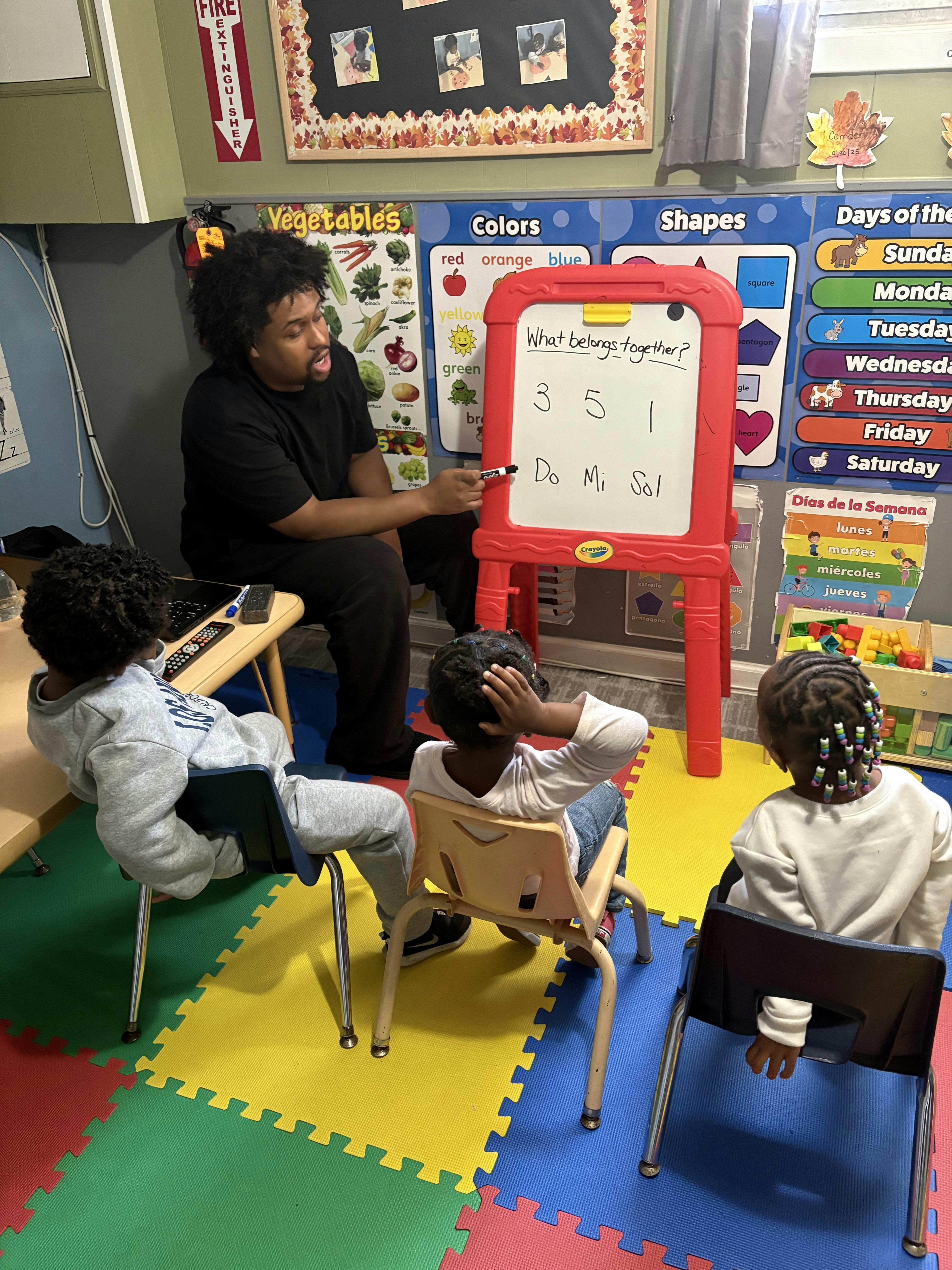 Person teaching three children in a classroom with a whiteboard that says "What belongs together?"