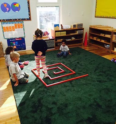 Children in a classroom with one child standing in a red spiral on a green rug