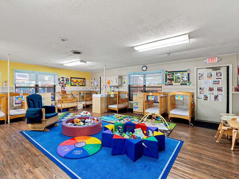 Classroom with multiple infant cribs, blue rug, and play area with toys