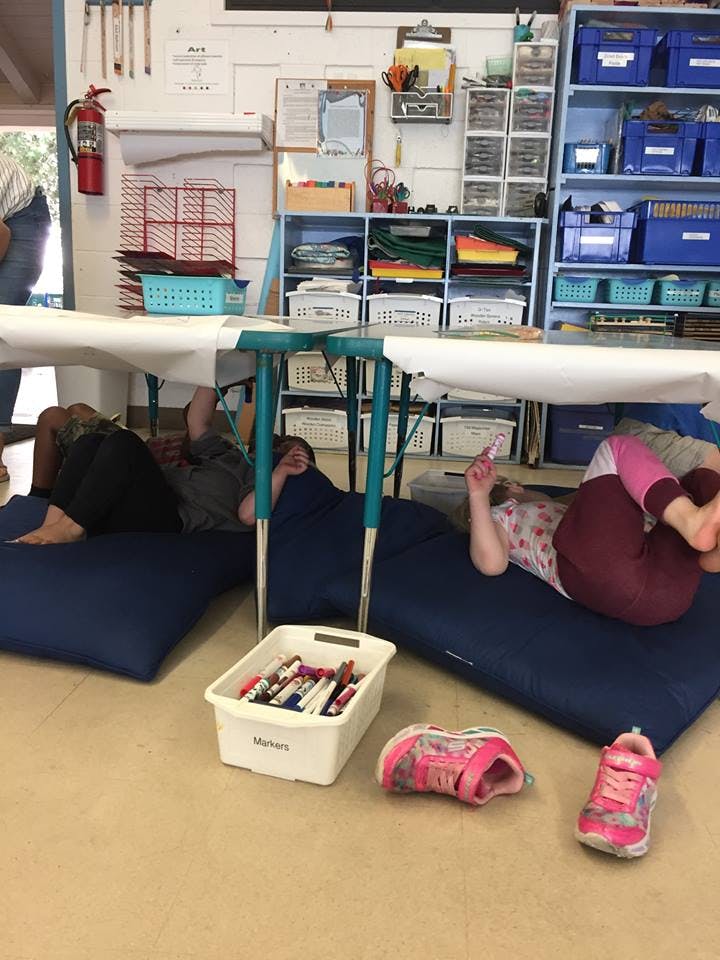 Children lying on blue cushions under a table in a classroom