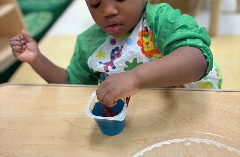 Child in a green smock and bib dipping a stick into blue liquid in a container