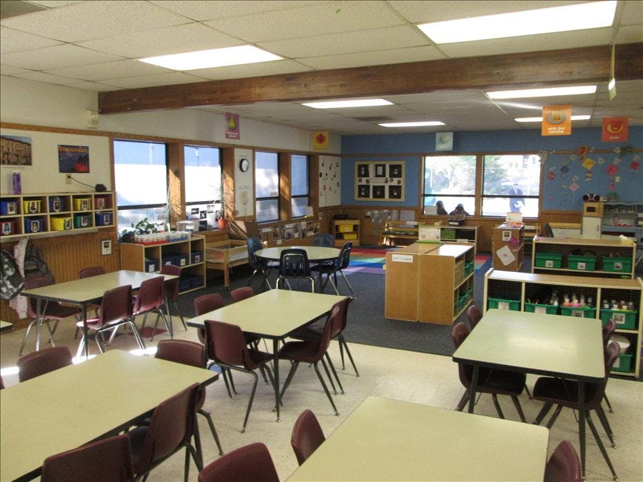 Empty classroom with multiple tables and chairs, storage units, and large windows