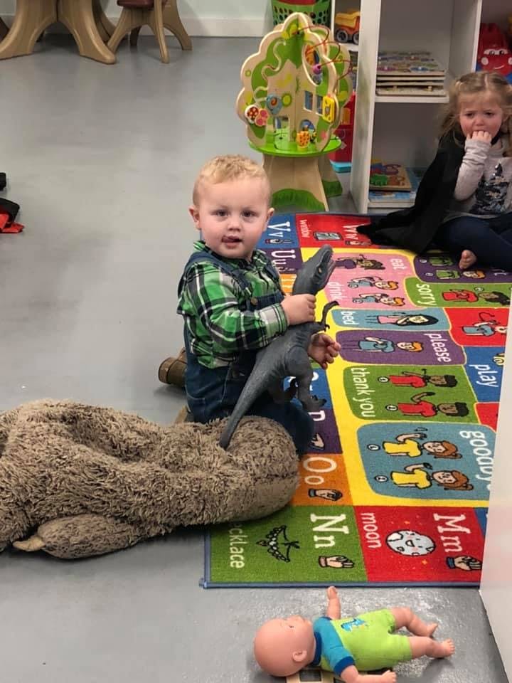 Child sitting on floor holding a dinosaur toy, another child in background