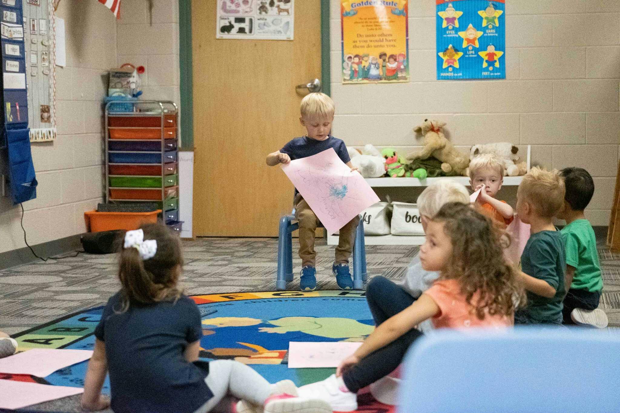 Child standing on a chair holding up a piece of paper to other children sitting on a rug