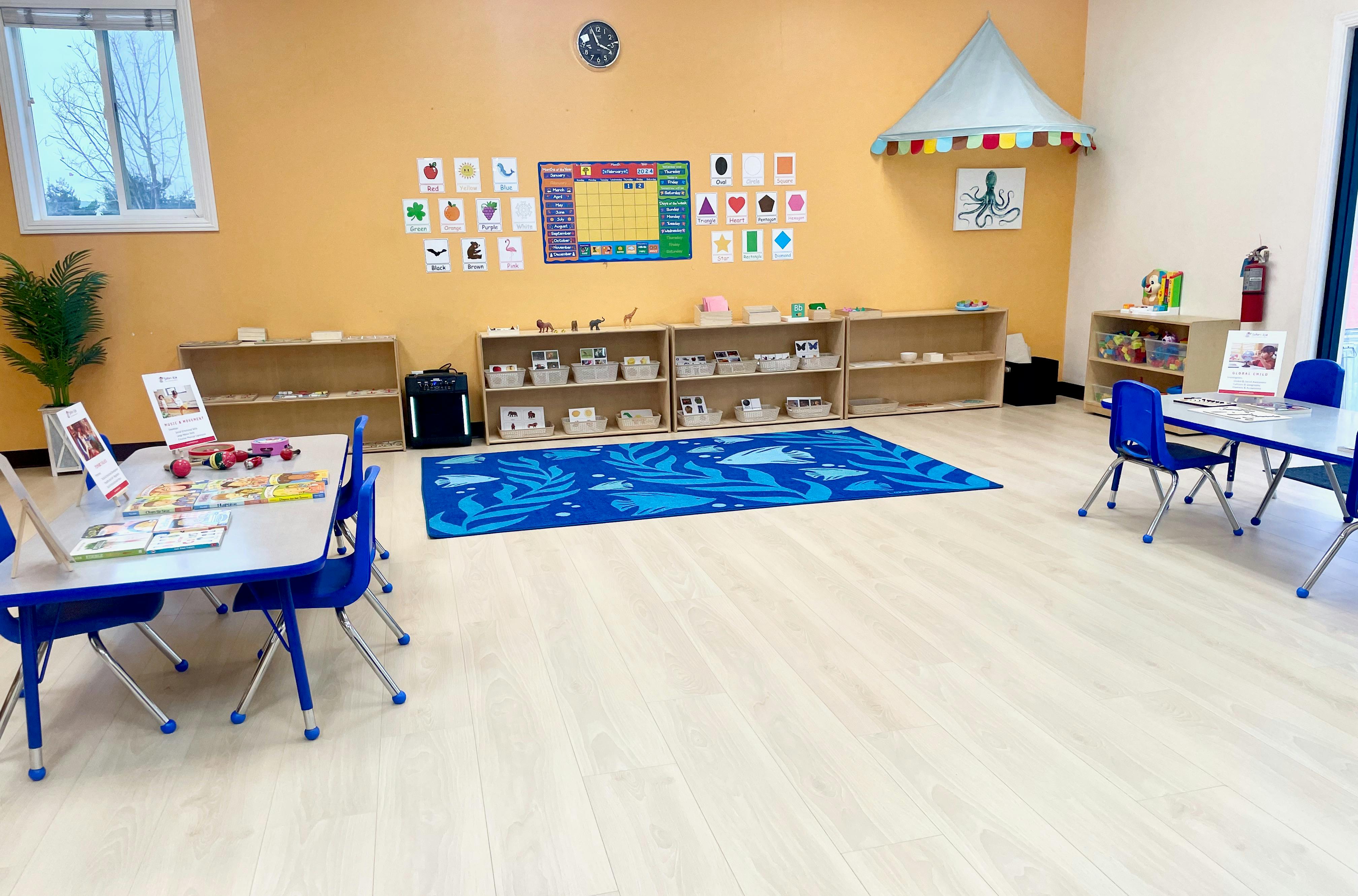 Classroom with small tables and chairs, shelves with learning materials, and a blue rug