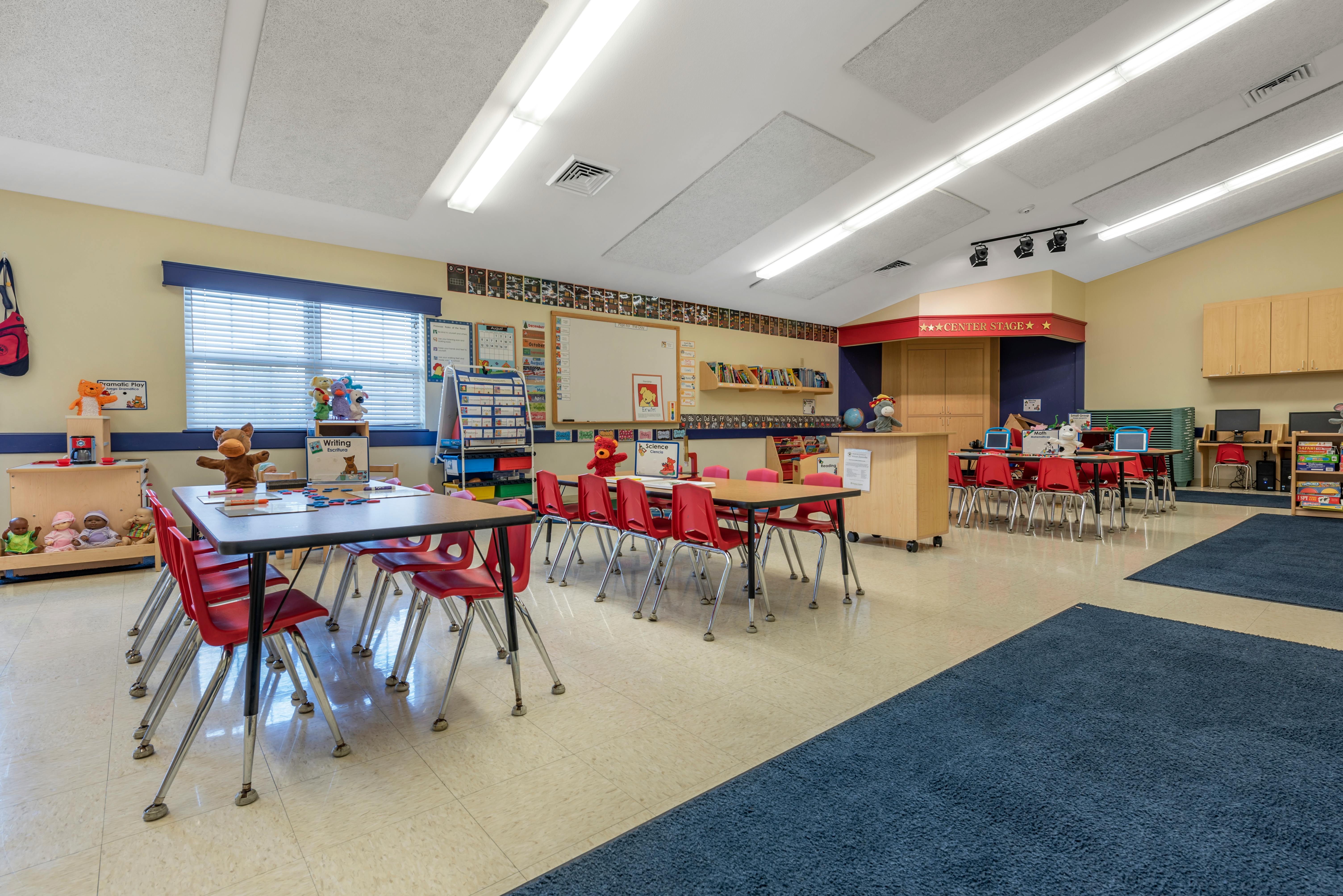 An empty classroom with several small tables, red chairs, and learning materials