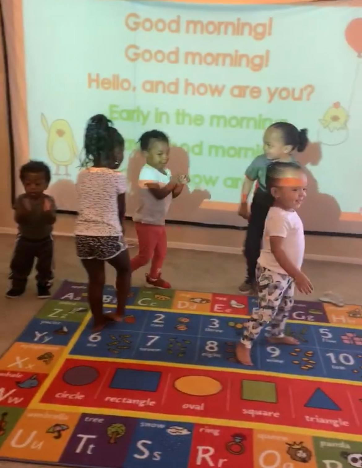Children standing on an alphabet and numbers rug with a projection on the wall